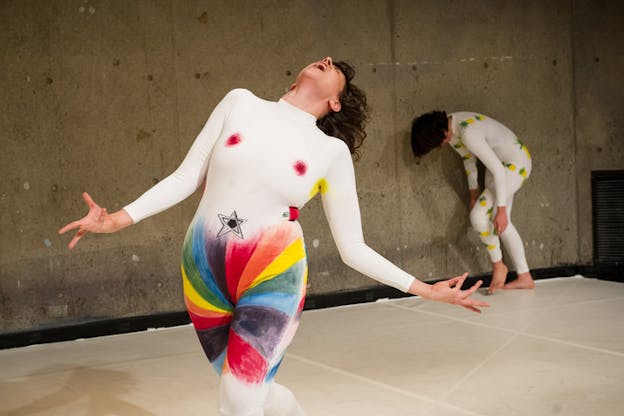 A performer dressed in a white leotard with rainbow swirl at the bottom of it and colorful stains on the breast and armpit areas holds their hands open as they scream with their head thrown backwards. Behind them another performer with a white leotard adorned with pineapples crouches as they are supported by the wall. 