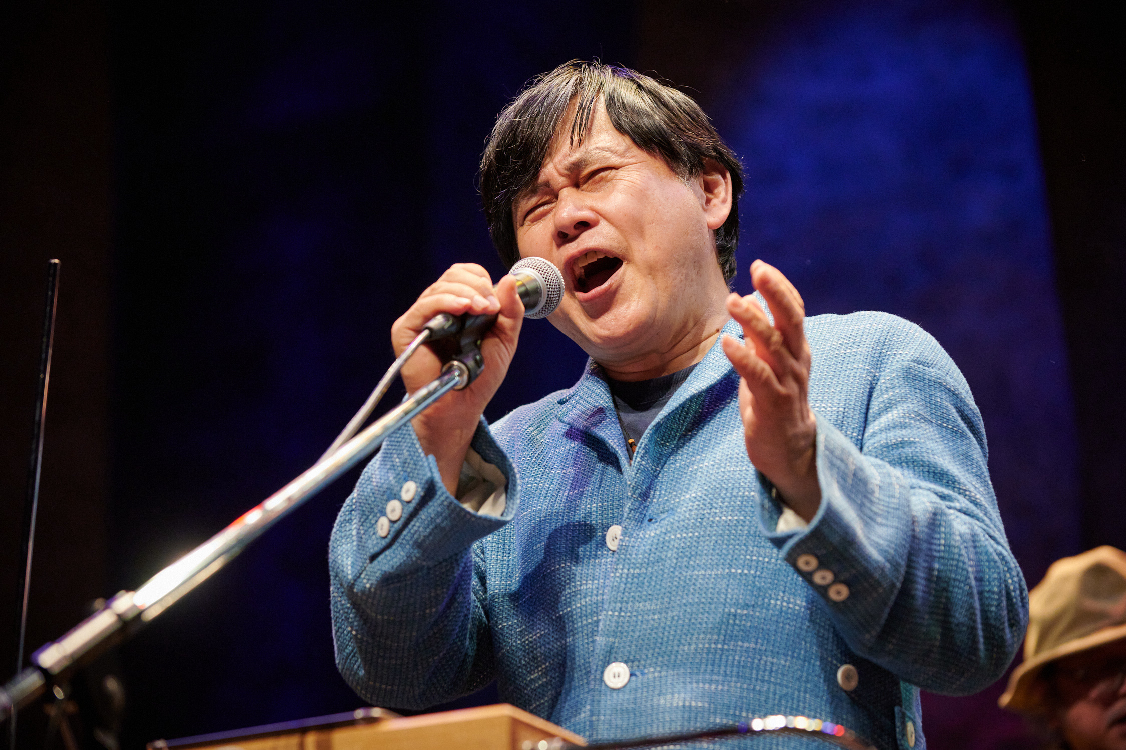 Koichi Makigami is photographed from below as he performs on a lit stage. He is visible from the waist up, turned slightly towards the left, standing behind a theremin and a microphone. His eyes are closed and his brows are furrowed as he vocalizes into the microphone which he holds with his right hand. His left hand is also raised at the same level. He is wearing a woven, light blue blazer with white buttons. 