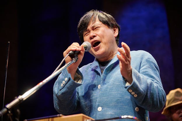 Koichi Makigami is photographed from below as he performs on a lit stage. He is visible from the waist up, turned slightly towards the left, standing behind a theremin and a microphone. His eyes are closed and his brows are furrowed as he vocalizes into the microphone which he holds with his right hand. His left hand is also raised at the same level. He is wearing a woven, light blue blazer with white buttons. 
