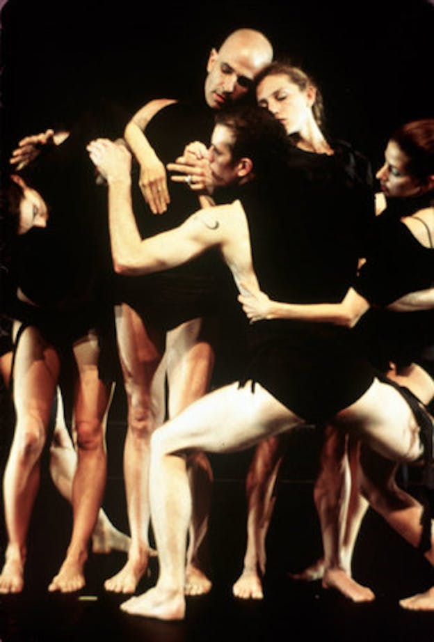 Several performers wearing black shirts and black underwear are intertwined within a dark space.The performer hold each other and close their eyes. 