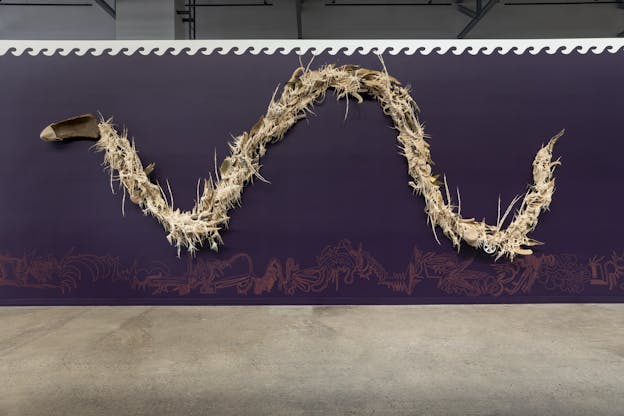 A bone-colored sculpture is mounted against a purple wall with linear orange illustrations along the bottom. The sculpture is a long serpentine trail that begins with a head-like piece of wood on the left side and extends in a wave to the right. The body is composed of off white, stringy spiked fringe wrapped around sticks and leaves, among other found objects including shells, a plastic toy wheel, and beer bottles. Small toy birds are nestled within the body. 