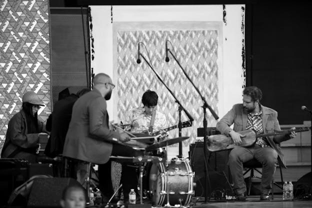 Black and white photograph of performers playing musical instruments such as guitars and drums in front of geometrical patterned tapestries. Two microphones stand on the front of them.  