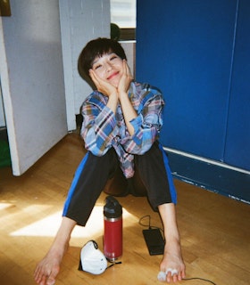 Hsiao-Jou sits on the wooden floor in the corner of a rehearsal space with an open white door behind her and blue padded panel walls to her right. Her body faces the camera, knees are up with her bare feet in front of her, her elbows are braced on her knees and her hands cradle her face on each side. Face tilted slightly up, she smiles into the camera. She wears a blue, brown and pink plaid button up shirt and black sweatpants with blue racing stripes.