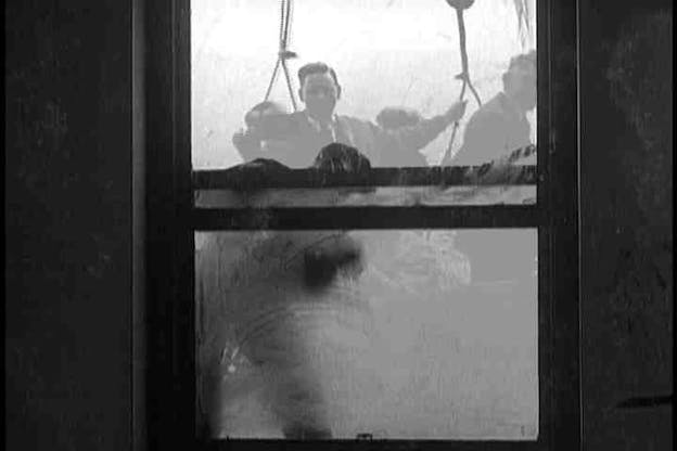 A black and white video still from the perspective inside of a building as people outside of it wash its window.