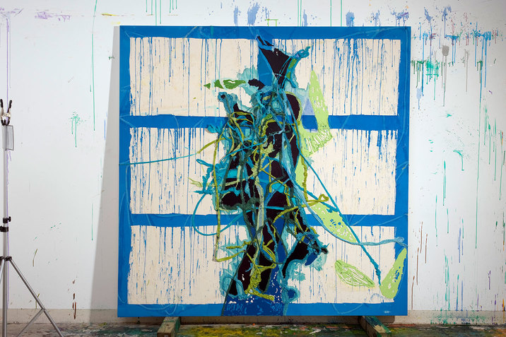 Installation view of a canvas divided in three white rectangles thickly lined with sapphire blue and dripping paint that extends to the wall beyond the canvas. In the center is an abstract accumulation of varying shades of blues and greens. 