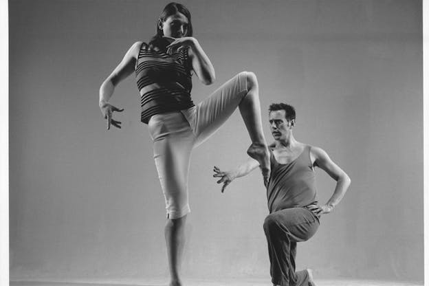 Image of performer supporting their weight on one leg as the other is angularly lifted to their side. Behind them another performer is kneeling on one leg and reaching towards something beyond the image view. 