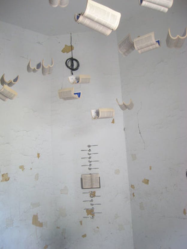 An installation image of a white room with the paint chipping off the walls. Opened paperback books hang suspended from the ceiling. On the far wall, one open book hangs with spoons above and below it. 