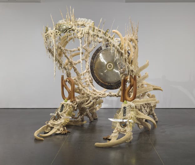 A sculpture, made primarily from beige, tan, and off white organic materials, in the form of an archway or alcove with a metal gong hanging in the center. The frame is coated in plastic and fabric with a rough, irregular texture and elongated spikes extending from the top. A curved portion hangs over, resembling the shape of a large upper and lower jaw. Long stalks of loofah line the edges, appearing tendril-like. Ornamental, smooth carved pieces of wood decorate either side of the arch, and transparent, teardrop-shaped orbs hang from the arch. Centered at the apex of the arch is a crab made from nacre or mother of pearl. Two daggers carved from mother of pearl point inwards towards each other on either side of the foot of the arch. 