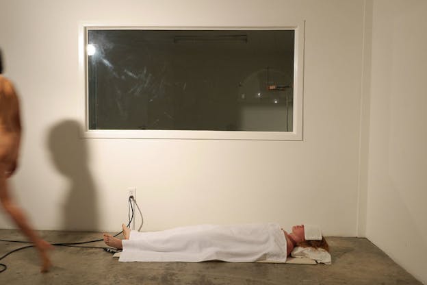 A figure lies on the floor with a white pillow underneath their head and a white towel covering their body and a smaller covering their eyes. Above them a glass window showing the inside of a room hosting a transparent dome with a bronze lamp inside it. 