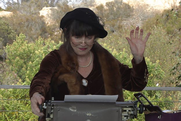 Person dressed in furs and pearls raises one palm and turns the knob on a typewriter against a backdrop of a hill covered in light and brown green foliage.