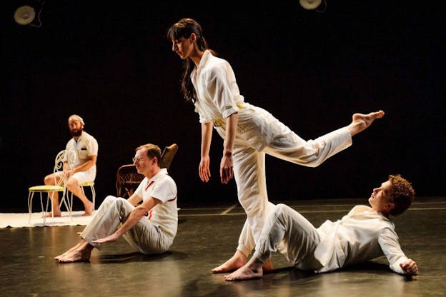Four performers dressed in white strike various poses on a dark lit stage: two lean back on the floor, one balances on one leg and leans forward with one leg bent behind them, and in the back corner, one sits on a chair. 