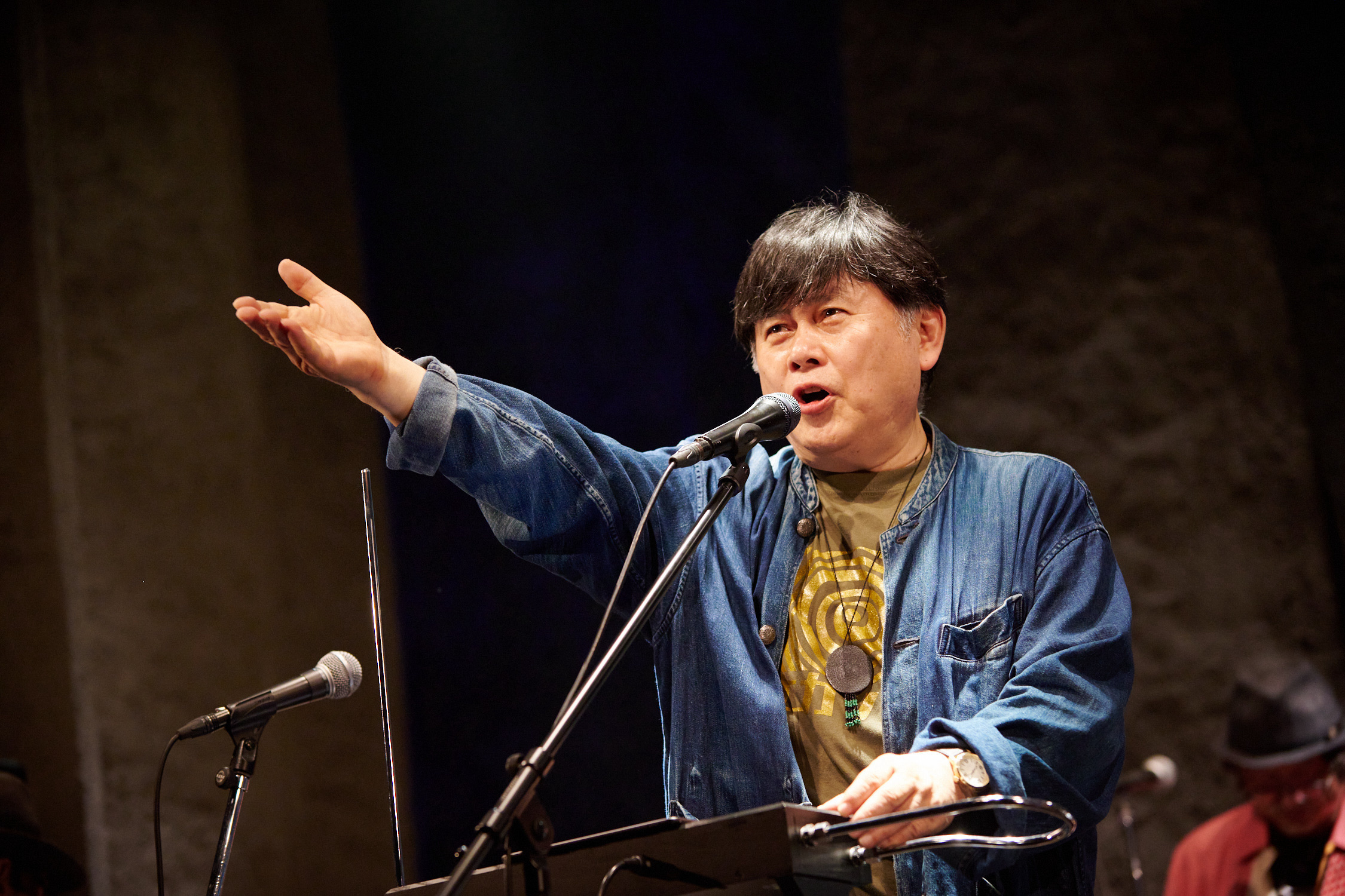 Koichi Makigami is centered on a lit stage. He is turned slightly towards the left, and standing behind a theremin and microphone. He looks outward as he vocalizes into the microphone, his left hand resting on the theremin and his right hand extended forward, palm turned up. He is wearing a denim collarless shirt with buttons, open over a tan graphic t-shirt, and a medallion necklace. 