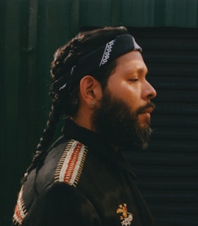 Guadalupe Maravilla stands in profile facing the right, before a dark gray metal garage door. His eyes gaze down and he wears a pair of beaded stud earrings, a black bandana folded and tied around his head, and a black jacket with embroidered details along the shoulder, back, and breast. 