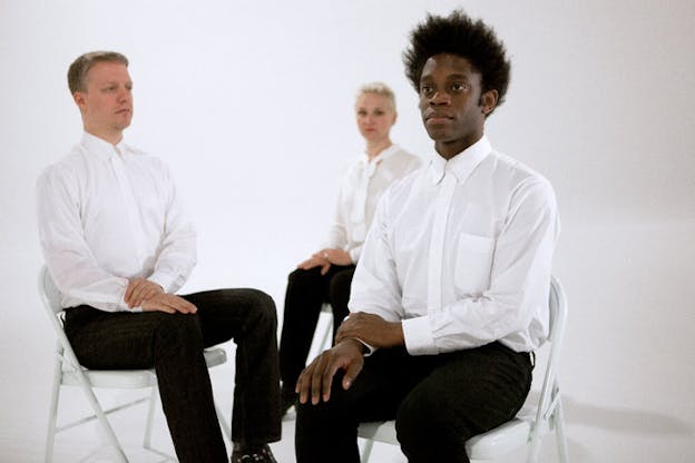 Three persons in an empty white room wearing white collared shirts and black pants, sitting on white chairs and staring straight ahead with blank expressions. They are positioned diagonally with the person closest in focus and the two behind blurred. 