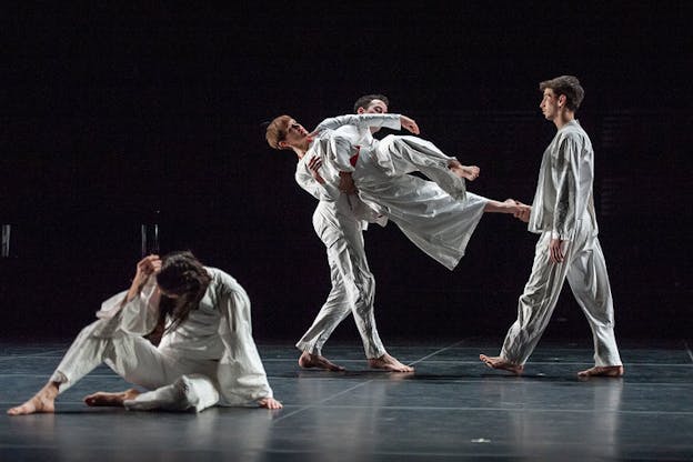 Performers wearing white tunic-clothing on a black stage with sharp lighting: two performers hold one performer by the performer's foot and waist while in front of them a performer sits on the floor and raises an arm towards their bent head. 