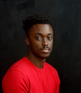 Nile Harris faces directly into the camera while his body is turned towards the right. He sits before a stark black backdrop and is dressed in a bright red shirt. 