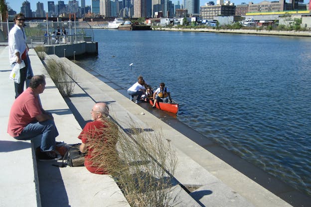 People sit in pairs on cement stairs leading to the water. A group of them on the last stair submerged in the water climb into a small red boat.