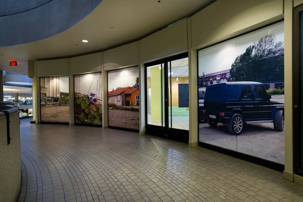 A curved wall displays screened photographs of a parked jeep, a fluorescent-lit room, a residential home, leaves growing beside a casette player, and a cracking parking lot of a concrete building with mountains in the distance. 