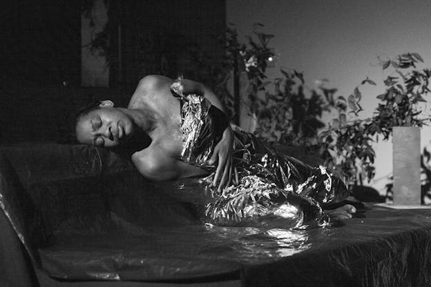 Black and white image of a person covered by a mettalic colored foil, laying on the ground. Their eyes are closed and their face is contorted in pain.