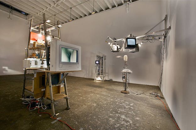Several sculptures are situated within an otherwise bare white room. On the left, a metal ladder supports various wires, pieces of cardboard, and a light bulb. A white television shows a blue image of part of a person. In the background, another wire structure supports a small black screen which projects something. On the far right, a metal structure attrached to the wall supports three screens which project images. 