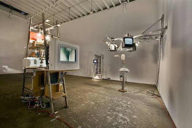 Several sculptures are situated within an otherwise bare white room. On the left, a metal ladder supports various wires, pieces of cardboard, and a light bulb. A white television shows a blue image of part of a person. In the background, another wire structure supports a small black screen which projects something. On the far right, a metal structure attrached to the wall supports three screens which project images. 