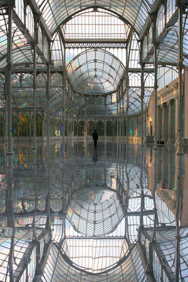 An image of a woman dressed all in black standing with her back to the camera within a large glass building. Light reflecting within the building creates small rainbows around the space. 