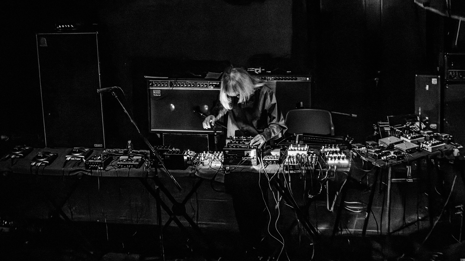 A black and white image of Keiji Haino bent over a long table covered in pedals, mixing boards, and other electronics. Behind him are several large amplifiers.
