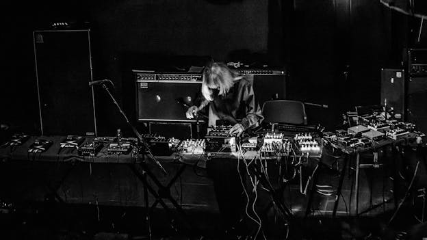 A black and white image of Keiji Haino bent over a long table covered in pedals, mixing boards, and other electronics. Behind him are several large amplifiers.