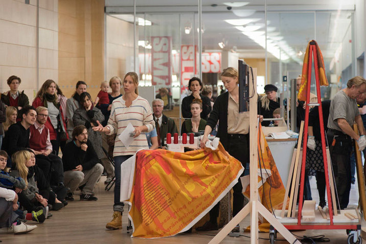 Two persons address an audience lined up against a wall, one holding a piece of paper and gesturing, while the other arranges a banner colored orange with illegible text draped over a table. 