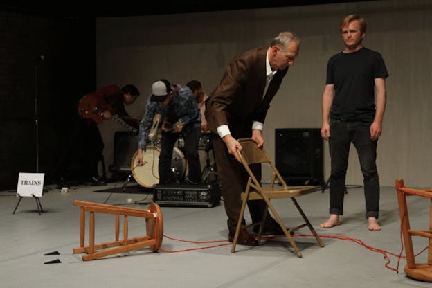 In the front right half of the frame a person in a brown suit leans over a plastic chair next to a person in a gray tee-shirt and jeans standing upright and two tipped wooden stools. In the bottom left three persons arrange musical instruments.