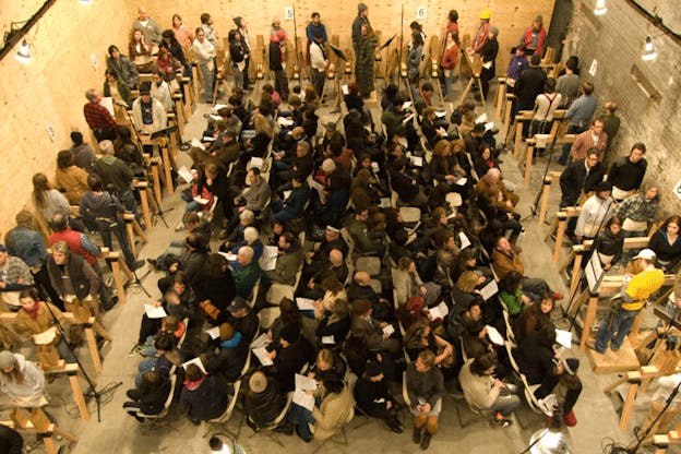 A performance still of several people within a brightly lit room. Two walls are plywood and the third is grey brick. On the perimeter of the room, people stand behind plywood shelves with microphones interspered. In the center of the room, audience members sit in chairs arranged in a rectangle with half of them facing one direction and the other hald facing the opposite direction. 