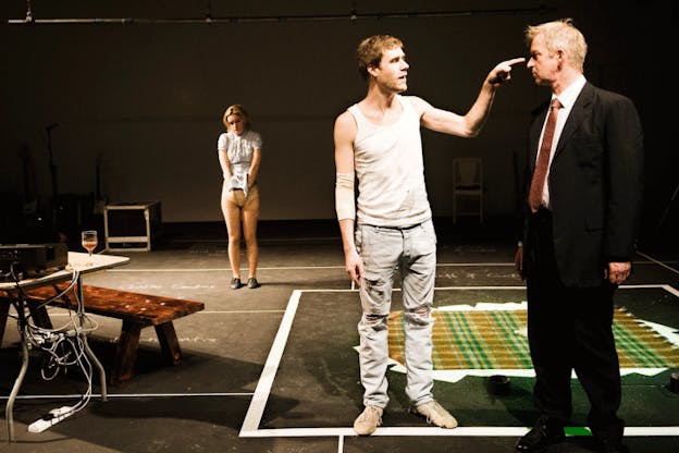 In the foreground, two performers looks intently at one another and one points to the forehead of the other. Behind them, there is a green plaid rug on the floor and another performer who looks distressed and stands hunched with their hands in their pants. 