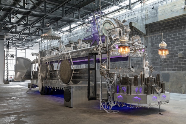 A large silver metalic sculpture build upon the skeleton of a schoolbus stands at three quarter view with the nose of the bus pointed to the right, in an industrial building with concrete floors, metal ceilings, and grey brick walls. The sculpture is intricately adorned with diamondtread metal sheets, various metal pieces, smaller sculptures, including table silverware, pots and pans, and clay idols in the forms of ears of corn. The top of the bus holds purple light fixtures and large metal sculptures including a winged dragon and a number of horses, escaped from a carousel that rests on the back end of the bus roof. Towards the front of the roof of the bus is a purple woven chair with a tall back made of spiked framing.