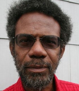 A close-up portrait of Pope L William wearing thin wire-rimmed oval sunglasses and a red shirt, standing in front of a white background. He looks directly at the camera. 