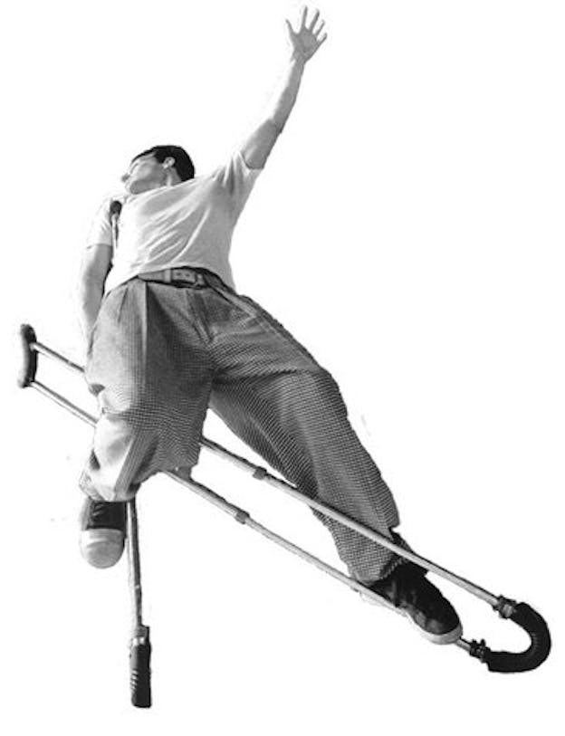 Bill Shannon leans back against a crutch with his right arm while his left arm extends long in front of him. He hooks his left foot into the other crutch which is diagonal from the ground. His image is against an all white background.