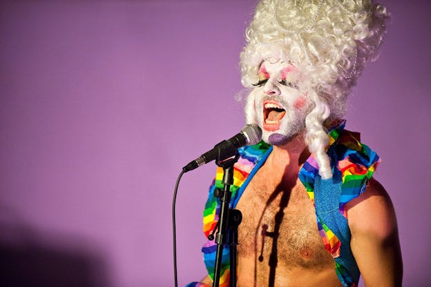 Close-up of a bare-chested performer wearing white face paint closing their thickly pink and gold rouged and fake eyelashed eyes while singing into a microphone, a beehive-shaped wig of platinum curls and pearls on their head and rainbow cloth wrapped around their shoulders. 