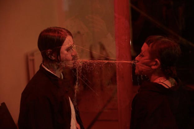 Two people facing each other in dim red lighting with thin cables on their faces and back of their heads that meet and tangle in the space between them as they pull apart.