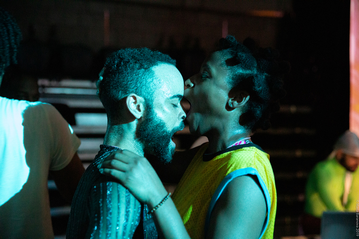 Two performers standing close face each other. The person in the left keeps their eyes closed as they speak, while the one on the right with an orange material in their mouth touches with it the forehead of the other and balances them with their hands on the other's shoulders.