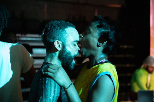Two performers standing close face each other. The person in the left keeps their eyes closed as they speak, while the one on the right with an orange material in their mouth touches with it the forehead of the other and balances them with their hands on the other's shoulders.