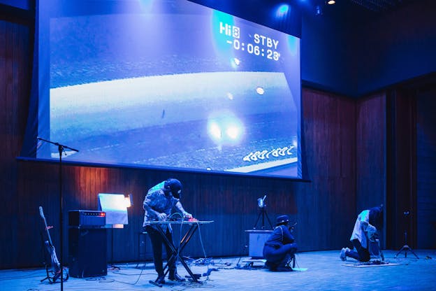 Three performers play on a stage washed in blue light, a projector screen lowered behind them. Nathan Young stands on the left side of the frame, bent over adjusting a soundboard. There is a guitar on a stand and two amplifiers stacked on top of one another to the left of where he stands. In the middle of the stage, Suzanne Kite crouches holding a walkie-talkie up to her face, her back facing the camera. Next to her, on the right side of the frame, Seth Cardinal Dodginghorse kneels holding their guitar, head tilted down to look at the pedal setup before them.