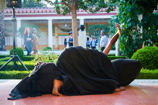 A performer completely covered in black material except their ends lays on top of a black sack on the floor.