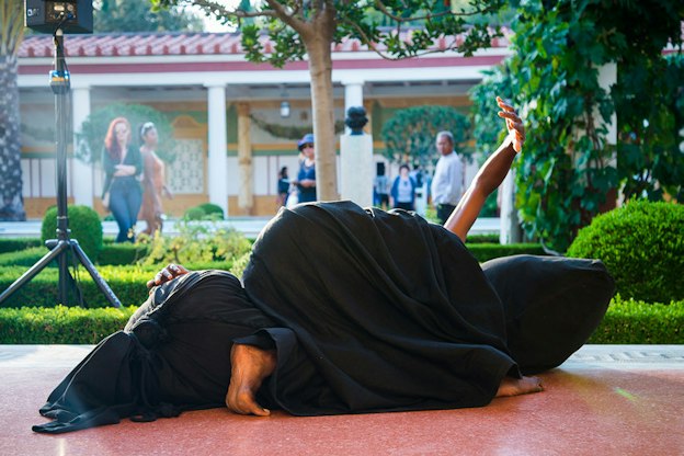 A performer completely covered in black material except their ends lays on top of a black sack on the floor.