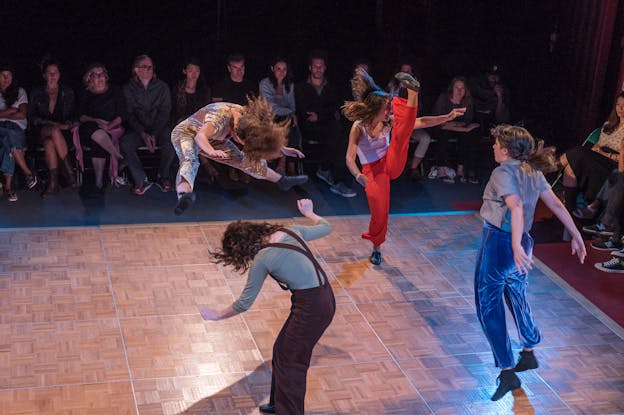 Four performers, one mid air with their legs elevated and the rest in various stages of jumping and lifting limps perform in a studio theater while a seated audience watches.