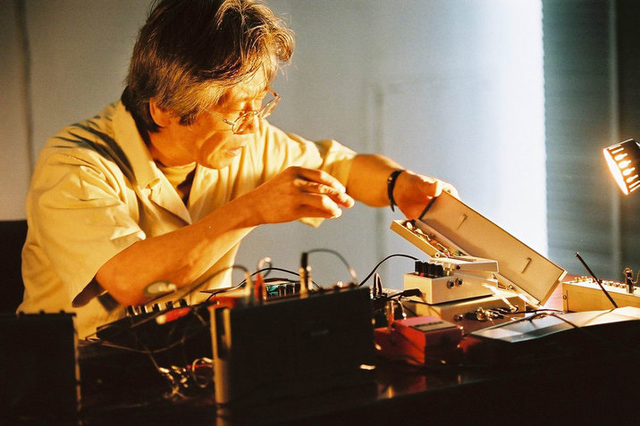 A figure illuminated by orange light sits behind a surface full of cables. They lift a white box towards themselves. 