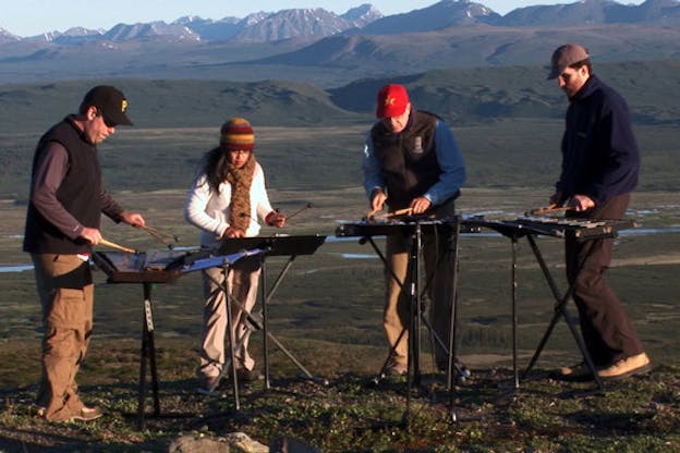 Four people dressed in heavily for the cold play xylophones. They are in a horizontal line, en face the camera. Mountains with peaks of snow and green plains stand behind them.