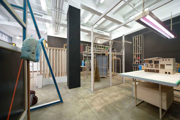 An installation image of numerous wooden structures within a black room. In the foreground, a fuzzy baby blue object rests on top of a wooden rod. To the right of that, a small model of a house sits atop a table. In the background, there are numerous glass paneled frames and a rack of beige clothing. 