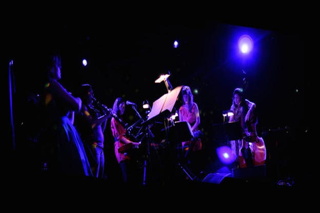 Low-angle shot of five performers standing in a semi-circle on a purple-lit stage playing various instruments including the cello and the clarinet. 