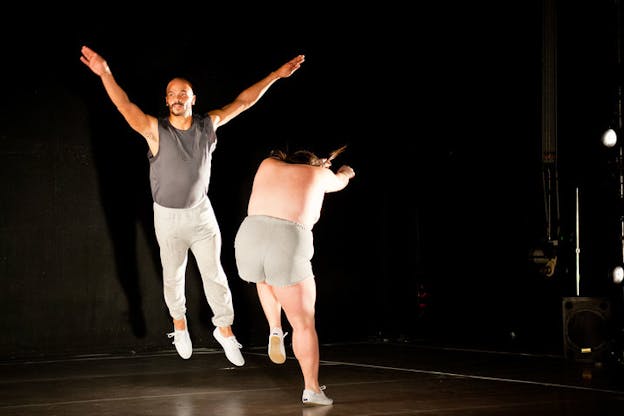 Two performers leap on stage: one facing forward and raising their arms in a V-shape while the other faces away and extends their arms outward with their head lowered. 