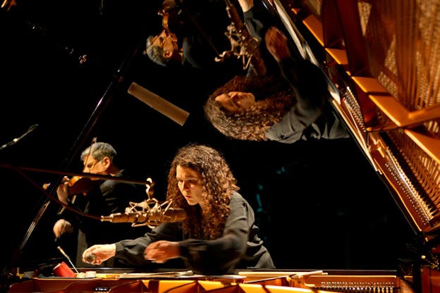 A person dressed in black behind a piano reaches towards its interior, a mircophone placed right in front of them. Behind her at the left a person with their side towards the viewer plays a violin. Both figures are reflected upside down on the right of the photograph on a glistering surface. 