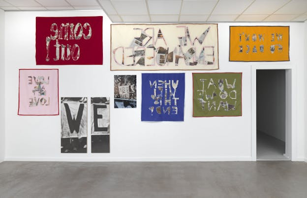 A gallery space with a white paneled ceiling and grey floors holds text-based visual arts works of different rectangular sizes framed and hung on a wall. Many works are made from fabric and feature a solid color background with backwards text constructed from collage. Phrases on the pieces include 